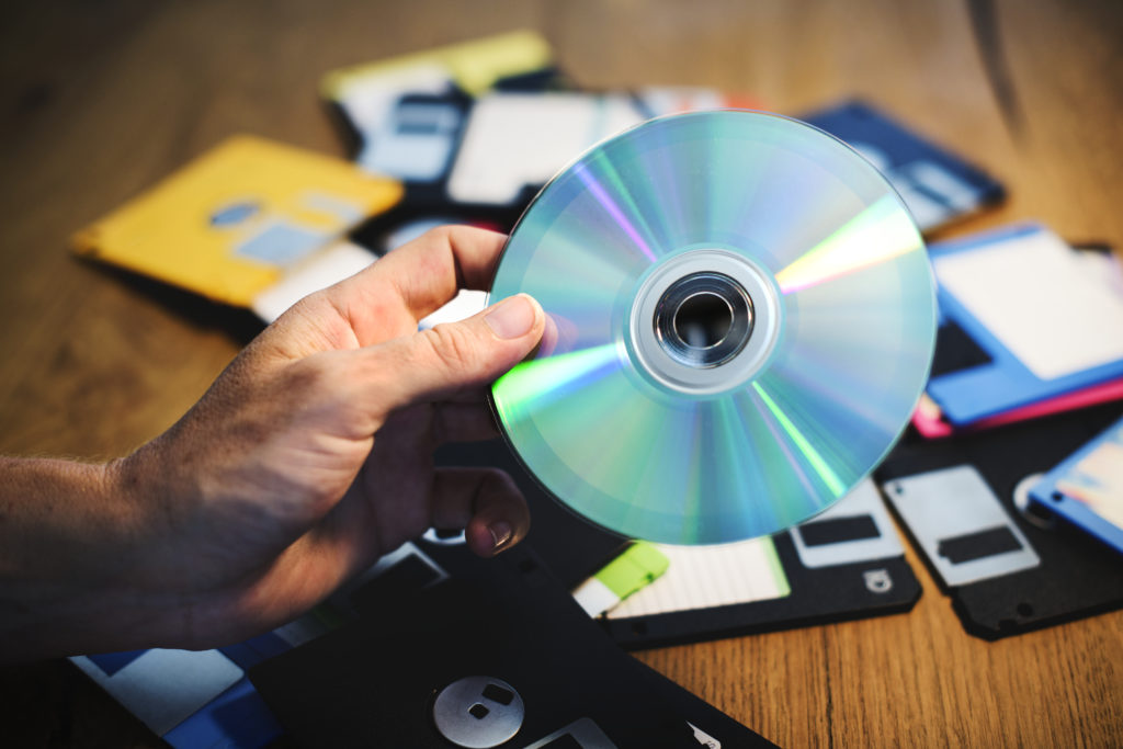 A person holding a CD ROM with a lot of floppy disks in the background